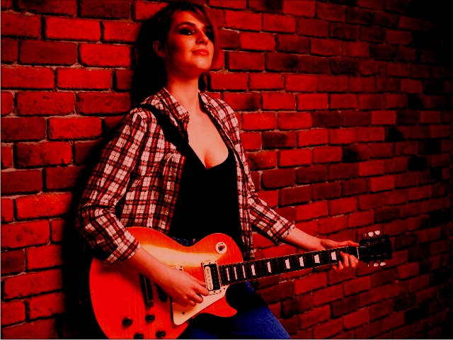 Woman holding an electric guitar against a red brick wall